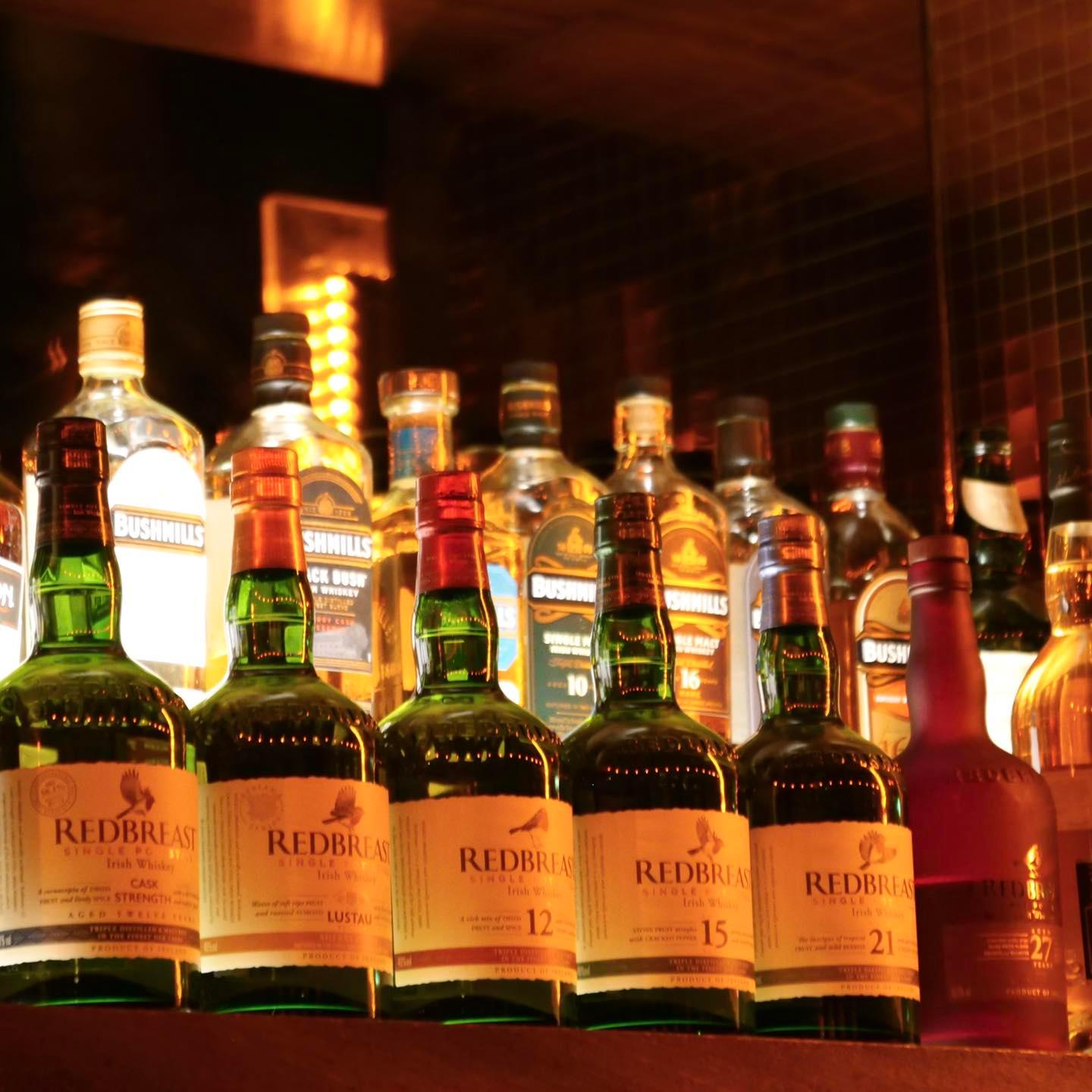 This photo shows a well-stocked shelf of Irish whiskey bottles, primarily featuring Redbreast, a highly regarded single pot still Irish whiskey brand. The bottles in the front row are labeled with different ages and types such as Cask Strength, Lustau, 12 Years, 15 Years, 21 Years, and 27 Years, showcasing a range of maturations and flavors. Behind them, you can also spot bottles of Bushmills, another famous Irish whiskey distillery, adding to the variety. The warm lighting creates an inviting atmosphere, typical of a cosy bar or whiskey lounge.