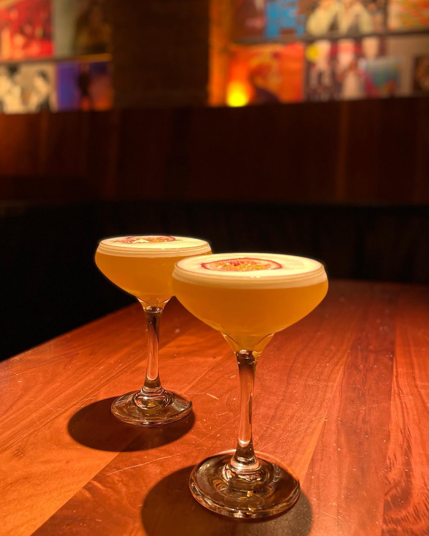 Two elegant cocktails in coupe glasses sit on a warm wooden table in a dimly lit bar. The drinks have a frothy top layer, each garnished with a slice of passion fruit. The background is softly blurred, featuring dark seating and colorful artwork on the walls, creating a cozy and inviting atmosphere.