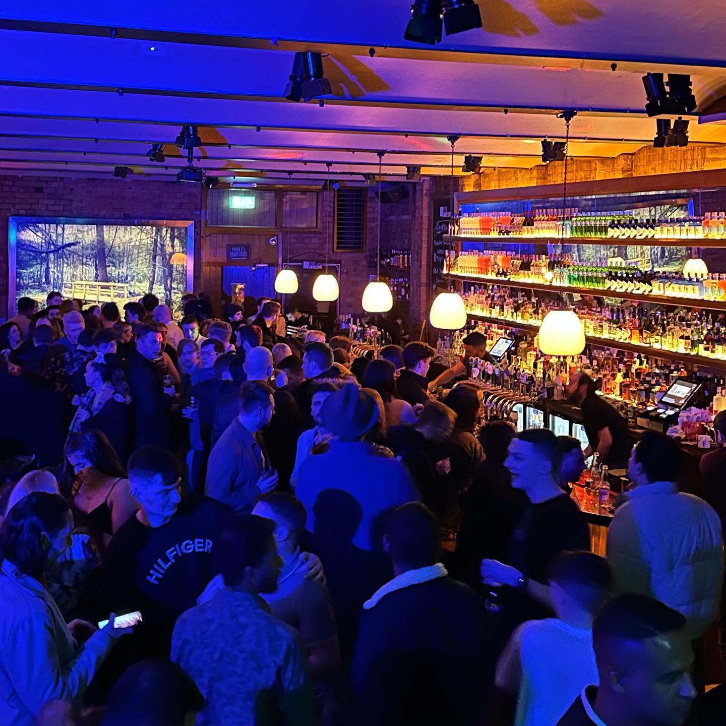 A vibrant bar scene packed with people enjoying a lively night out. The bar is warmly lit with hanging pendant lights above a long, fully stocked counter, while colorful ambient lighting in blue and orange bathes the crowd. Patrons are chatting, ordering drinks, and soaking in the energetic atmosphere. A digital screen on the back wall displays a scenic nature image, adding an artistic touch to the brick interior.