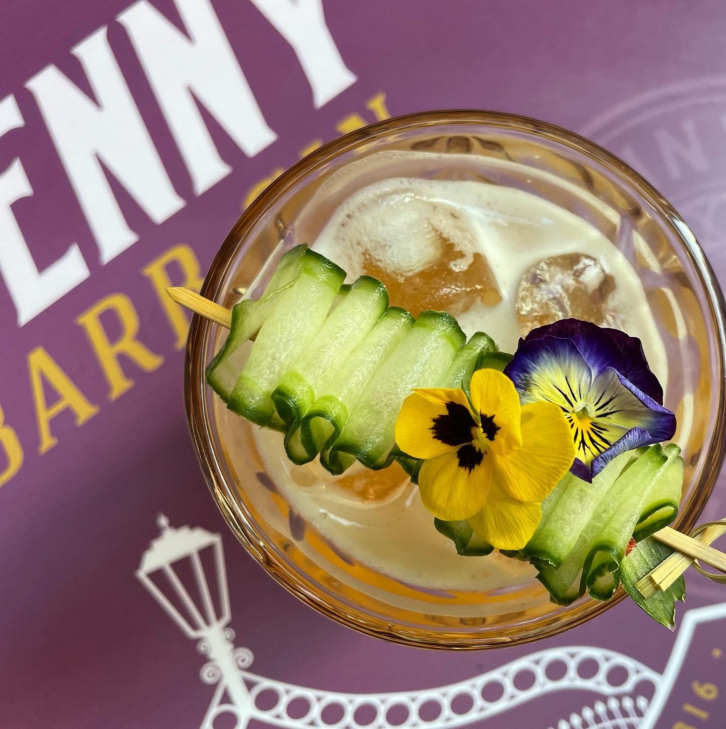 A top-down view of a creamy cocktail with ice cubes in a glass. The drink is garnished with a skewer holding intricately folded cucumber slices and two edible flowers, one yellow and one purple. The glass is placed on a purple surface featuring white and yellow text and graphics.