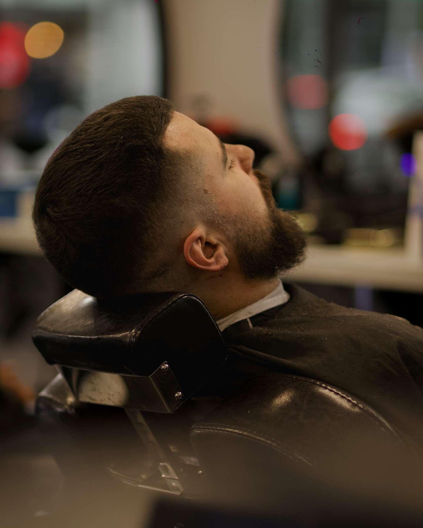A man with a fresh fade haircut and trimmed beard reclines in a barber chair, resting his head on the neck support,