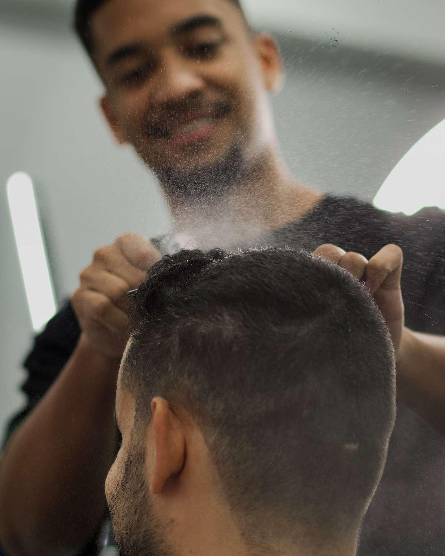Barber smiling while spraying water on a client's hair during a haircut, with the client's head seen from behind.