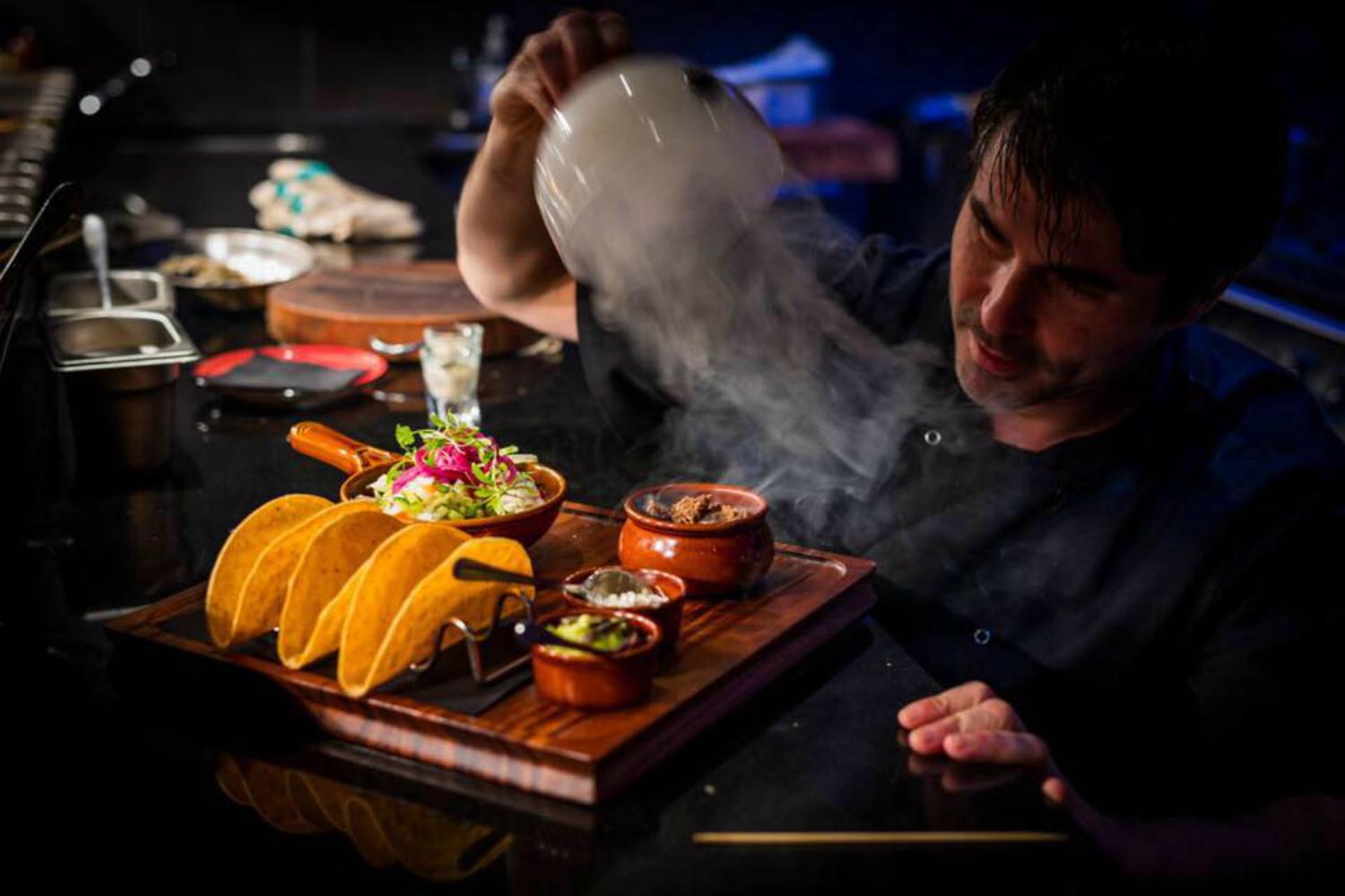 A chef in a dark kitchen reveals a smoking clay pot on a wooden tray, which also holds three upright hard-shell tacos in a metal stand, a bowl of fresh salad with pickled onions, and three small clay ramekins containing taco toppings such as guacamole, salsa, and sour cream. The scene is dimly lit, highlighting the vibrant colors and rising smoke.