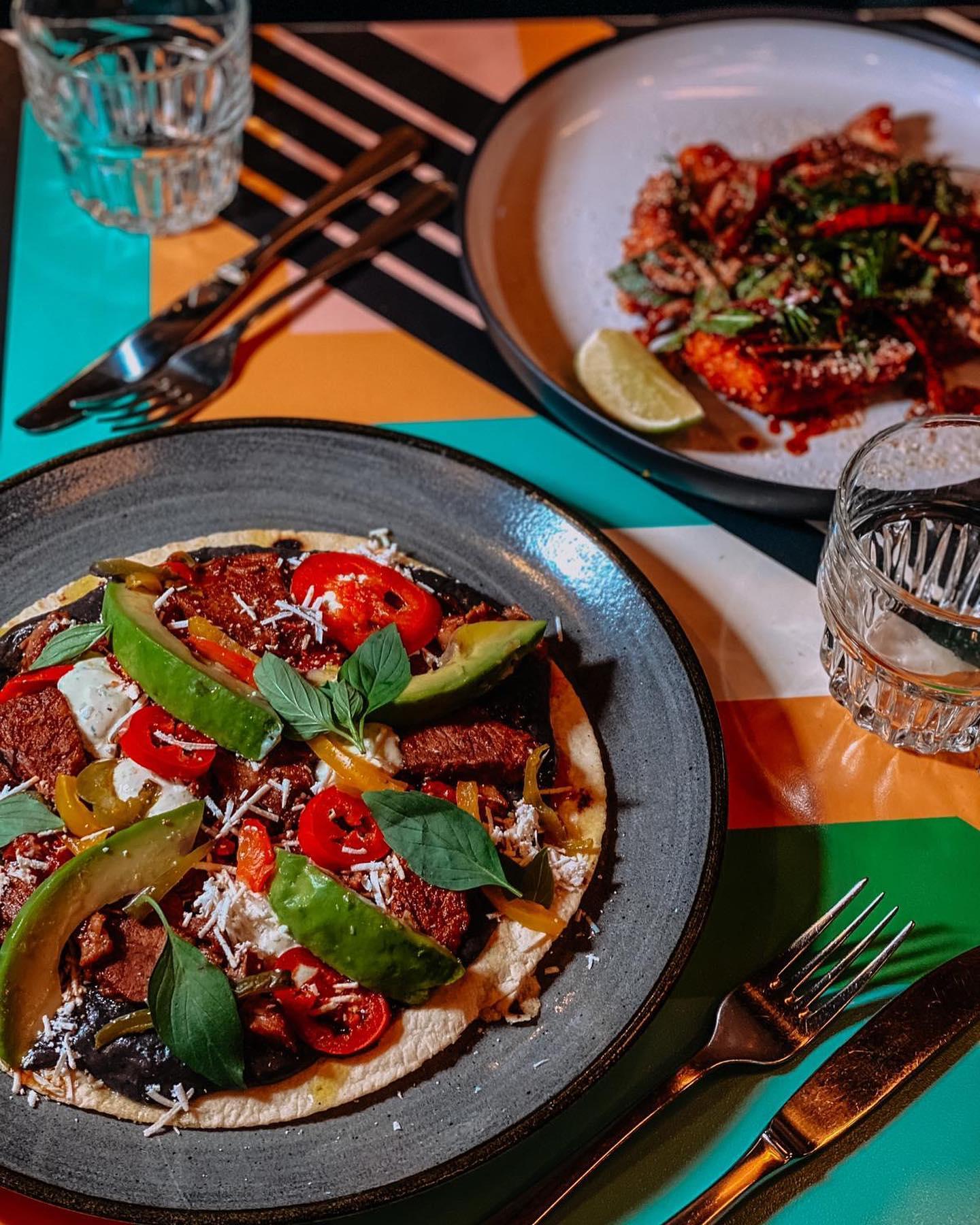 A colorful table setting with two plated Mexican dishes. In the foreground, a grey plate holds a tostada topped with grilled beef slices, avocado wedges, pickled peppers, cherry tomatoes, fresh basil leaves, crema, black beans, and grated cheese. In the background, a white plate features a saucy dish garnished with herbs, chili, and a lime wedge. Two glasses of water and cutlery are placed on a geometric-patterned table mat with teal, orange, black, and white colours