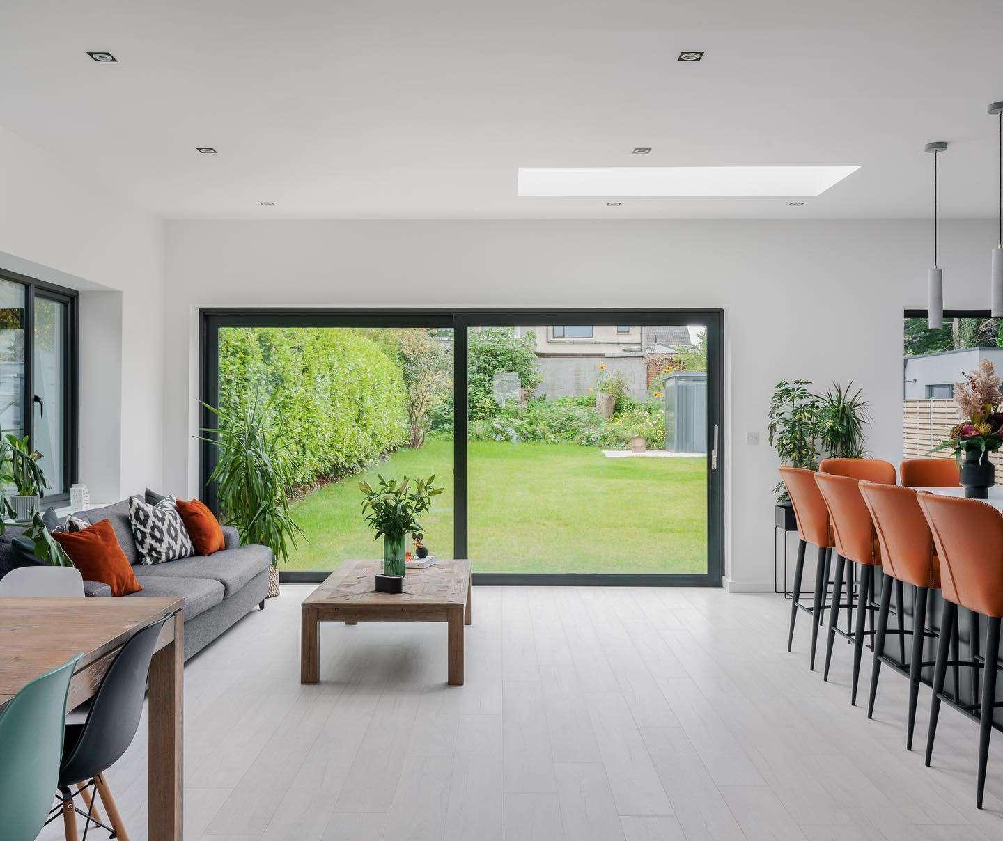 Modern open-plan living and dining area with large sliding glass doors leading to a green backyard. The room features a gray sofa with orange and black cushions, a wooden coffee table, and a dining island with orange bar stools. The space is bright with white walls, light wood flooring, and natural light coming from a skylight and large windows. Several indoor plants add greenery to the interior.