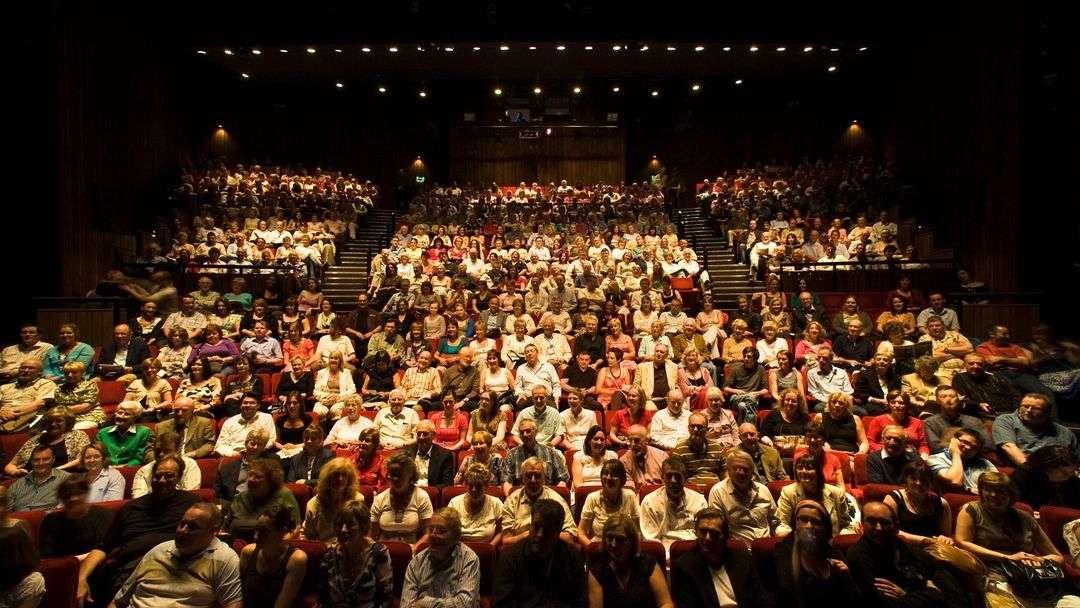 A large, diverse audience seated in a theater auditorium. The photo is taken from the stage, showing rows of people filling the seats, facing forward toward the stage. The theater is dimly lit with overhead lights focused on the crowd.