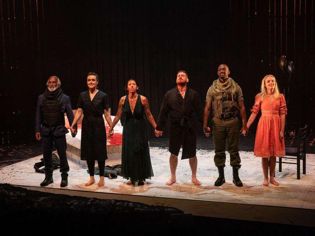 Six actors stand in a line on a stage, holding hands and taking a bow. The stage set includes a bed with a red and white blanket and a black chair. The actors wear a variety of costumes: the first man on the left is in a dark suit with a tactical vest and scarf; the next two women wear black dresses; the man in the middle wears a black robe and is barefoot; the next man wears military-style clothing and boots; the woman on the far right wears an orange dress and is barefoot. The background is dark with vertical lines of light.