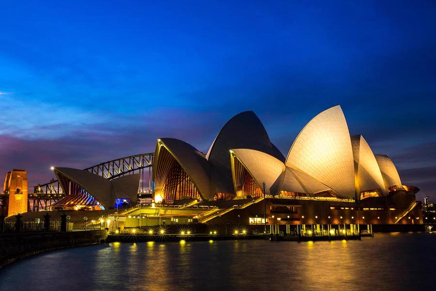 The Sydney Opera House illuminated at night with its iconic sail-like roofs, set against a deep blue sky and reflected in the harbor water, with the Sydney Harbour Bridge in the background.