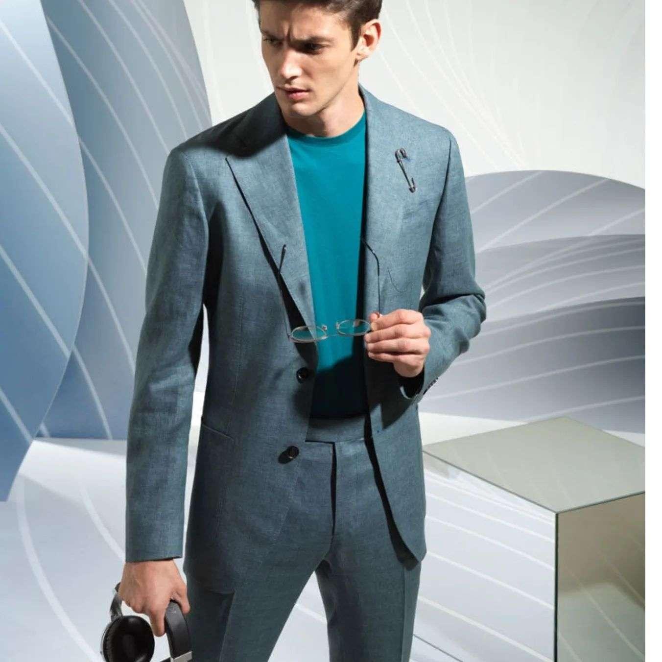 A man wearing a modern light blue suit with a teal shirt underneath, holding eyeglasses in one hand and headphones in the other, standing in front of a sleek, curved abstract background.