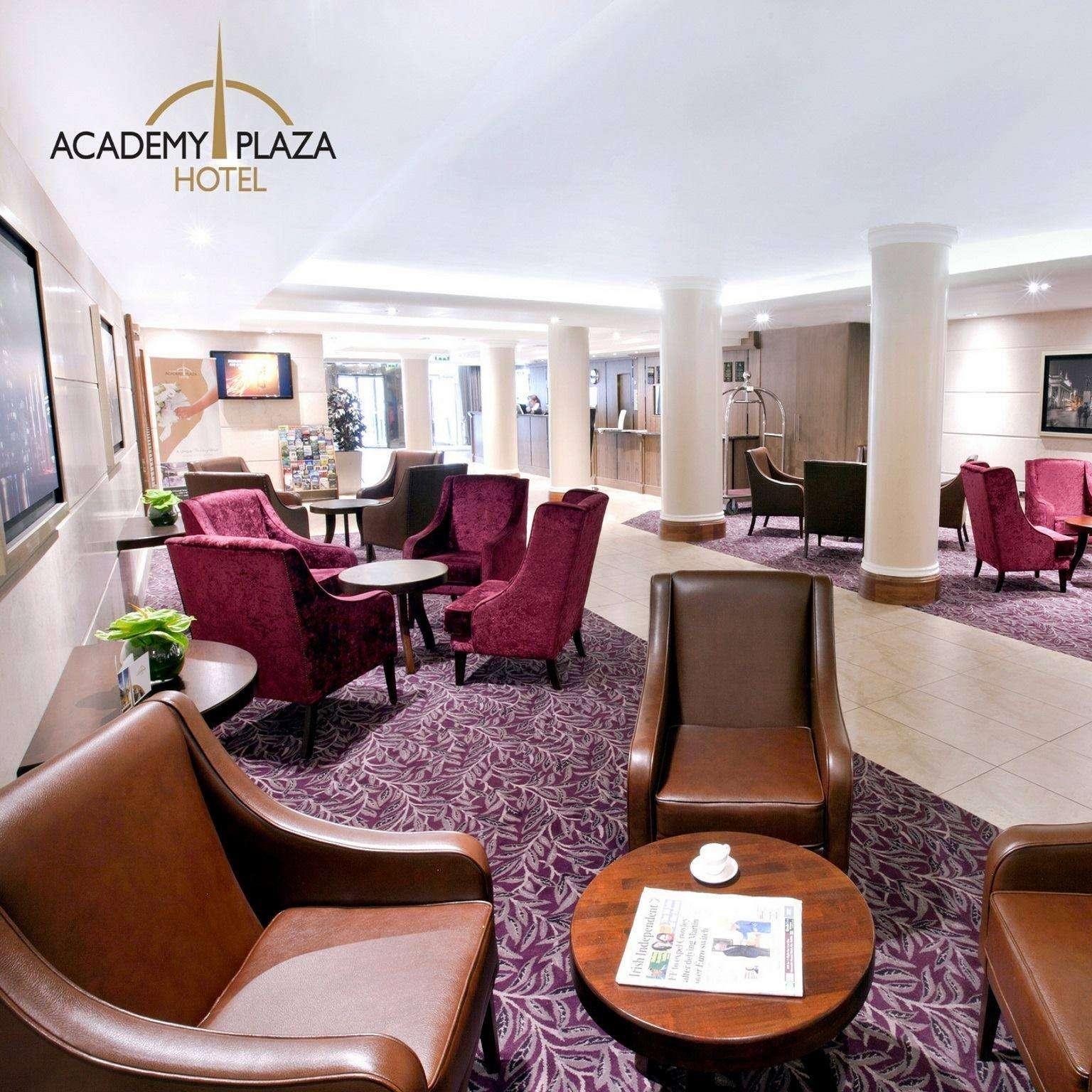 Bright and spacious hotel lounge with purple and brown armchairs arranged around wooden tables on a patterned purple carpet and cream tile floor; white columns and reception desk visible in the background; Academy Plaza Hotel logo in the top left corner.