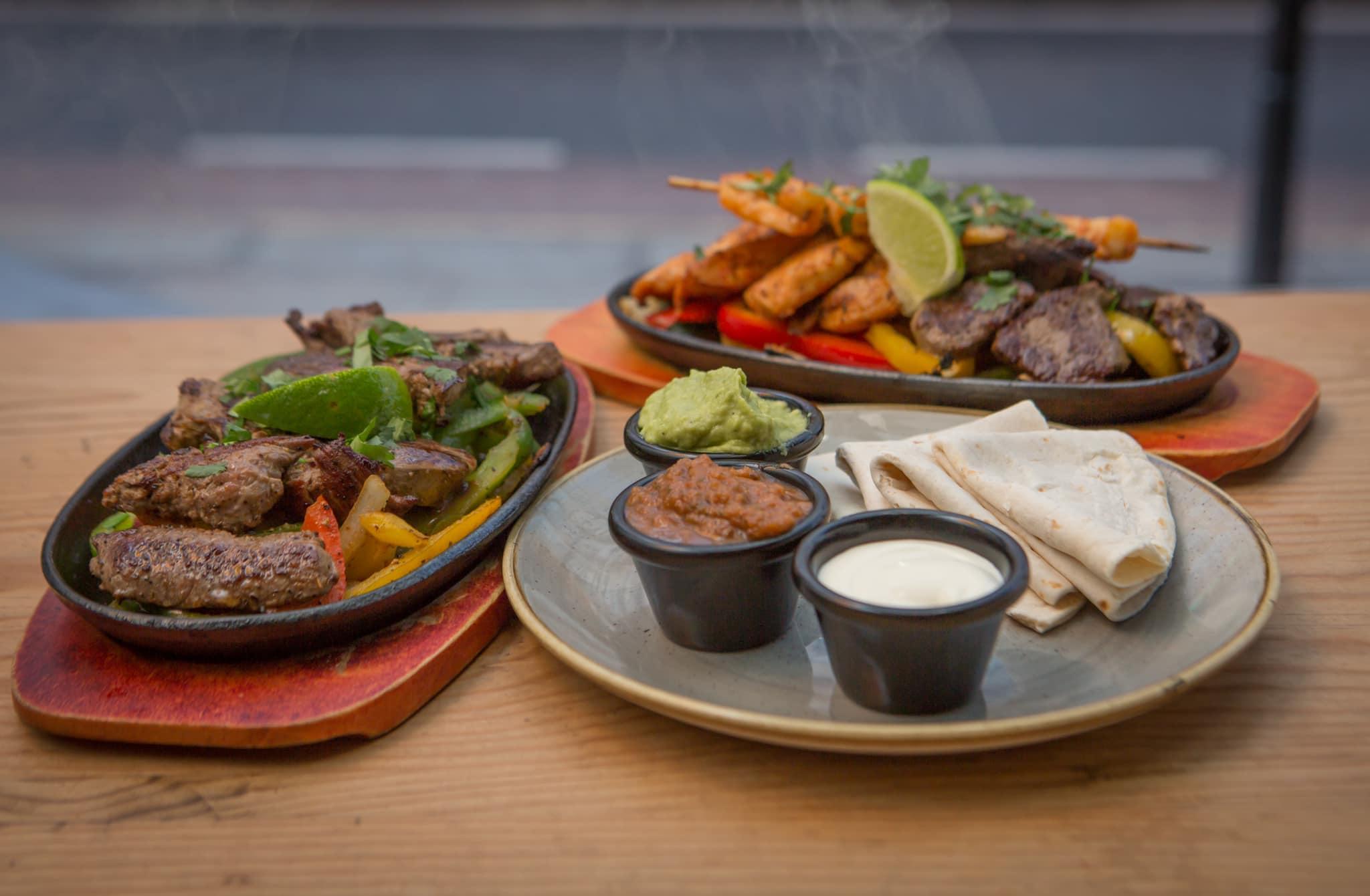A close-up image of a wooden table with three plates of food. On the left is a sizzling skillet with grilled beef strips and sautéed green, yellow, and red bell peppers, garnished with cilantro and a lime wedge. In the center is a round plate with three small black bowls containing guacamole, salsa, and sour cream, alongside folded soft flour tortillas. On the right is another sizzling skillet with grilled shrimp and beef strips on a bed of sautéed red and yellow bell peppers, garnished with fresh herbs and a lime wedge. The background is softly blurred.