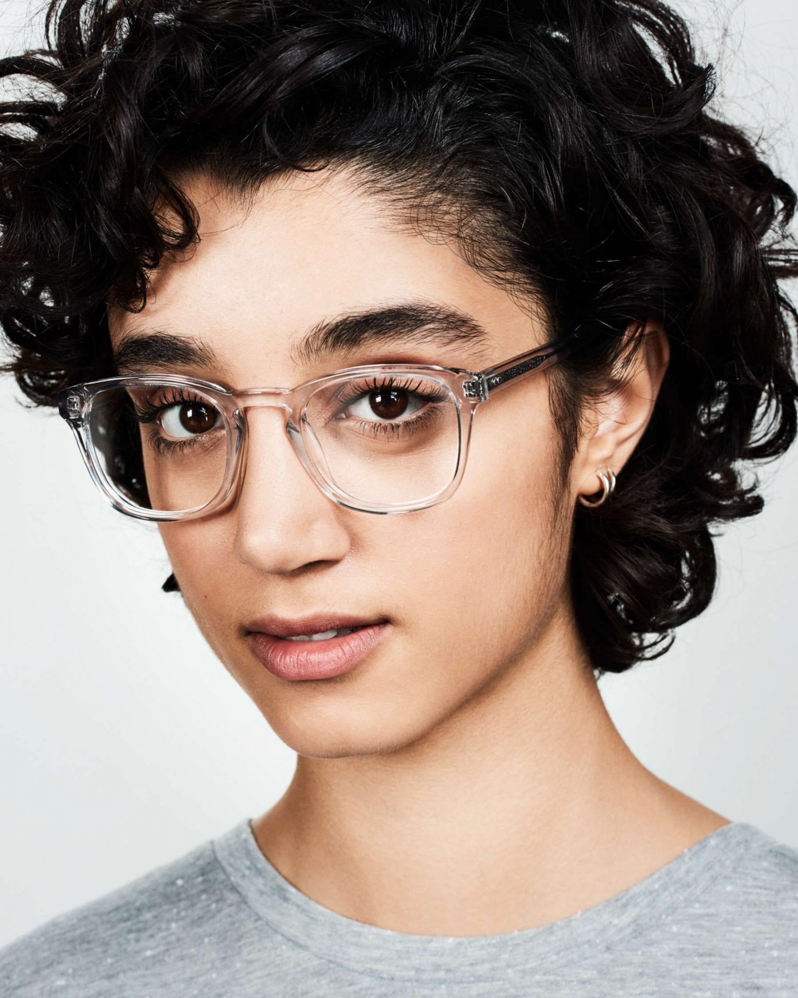 Close-up portrait of a person with short curly dark hair, wearing clear rectangular eyeglasses and a light gray shirt, looking directly at the camera with a neutral expression.