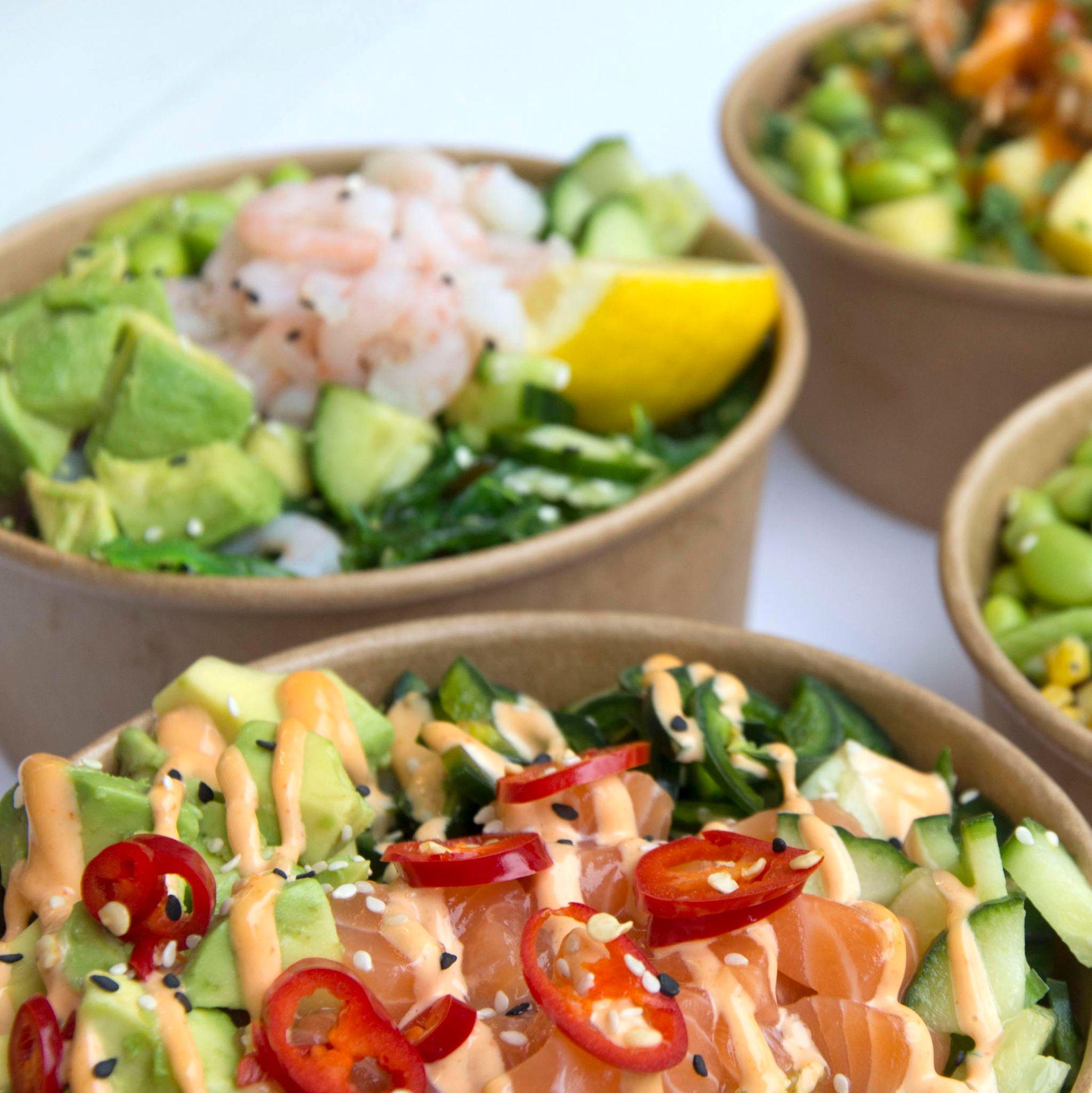 Close-up of assorted Hawaiian poké bowls in compostable containers, featuring diced salmon, avocado, cucumber, red chili slices, shrimp, lemon wedge, and a drizzle of spicy mayo on fresh greens.