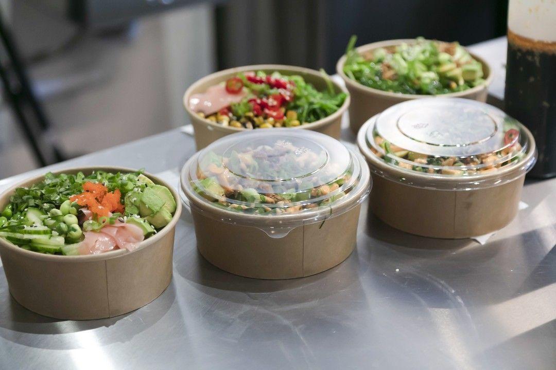 Five brown compostable bowls filled with assorted poké ingredients, including green vegetables, raw fish, and toppings. Some bowls are open and others sealed with clear plastic lids, displayed on a stainless steel counter.