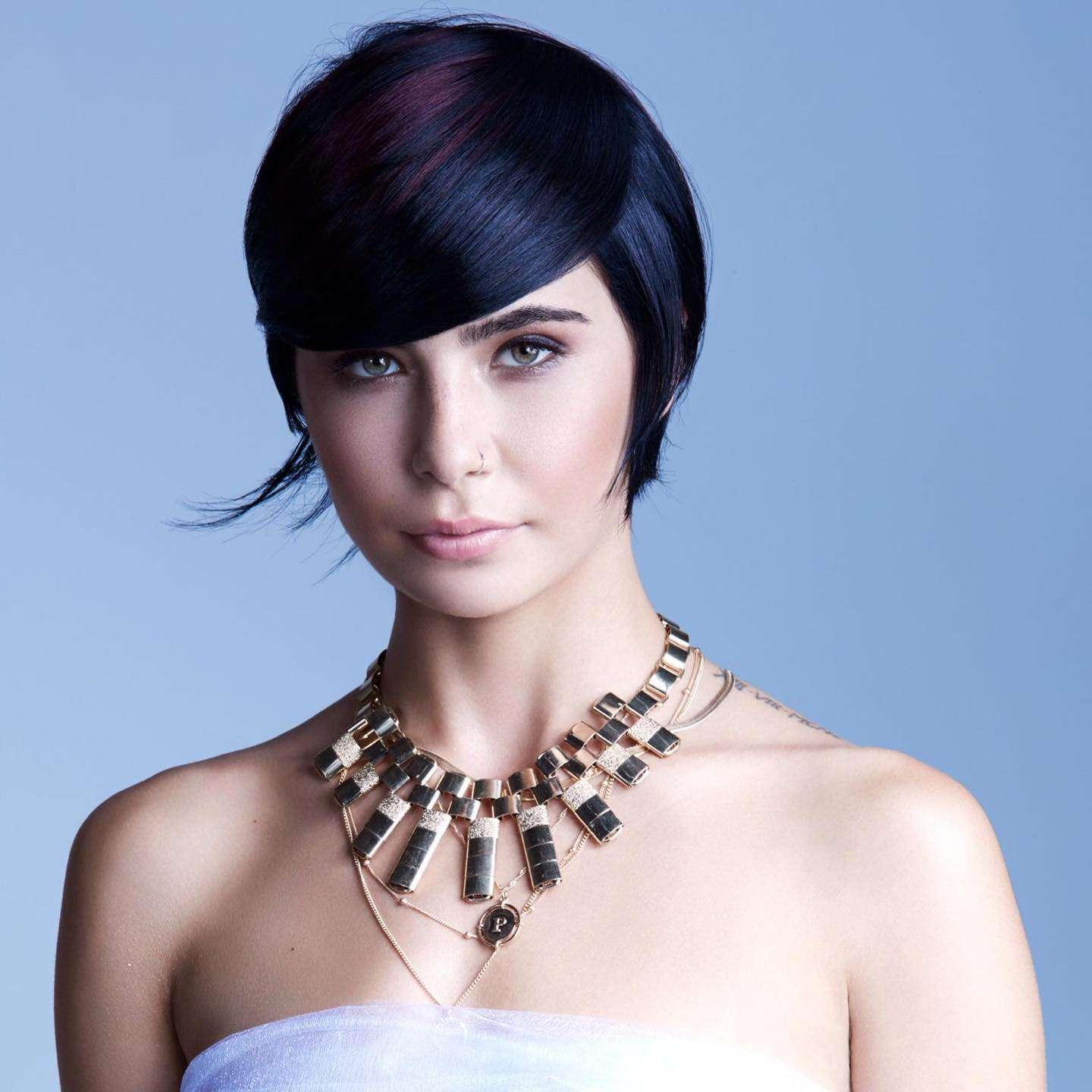 Woman with short, sleek black hair styled in a modern asymmetrical pixie cut, wearing a strapless white top and a bold, layered gold necklace. She has a light complexion, minimal makeup, and a nose ring, posed against a light blue background.