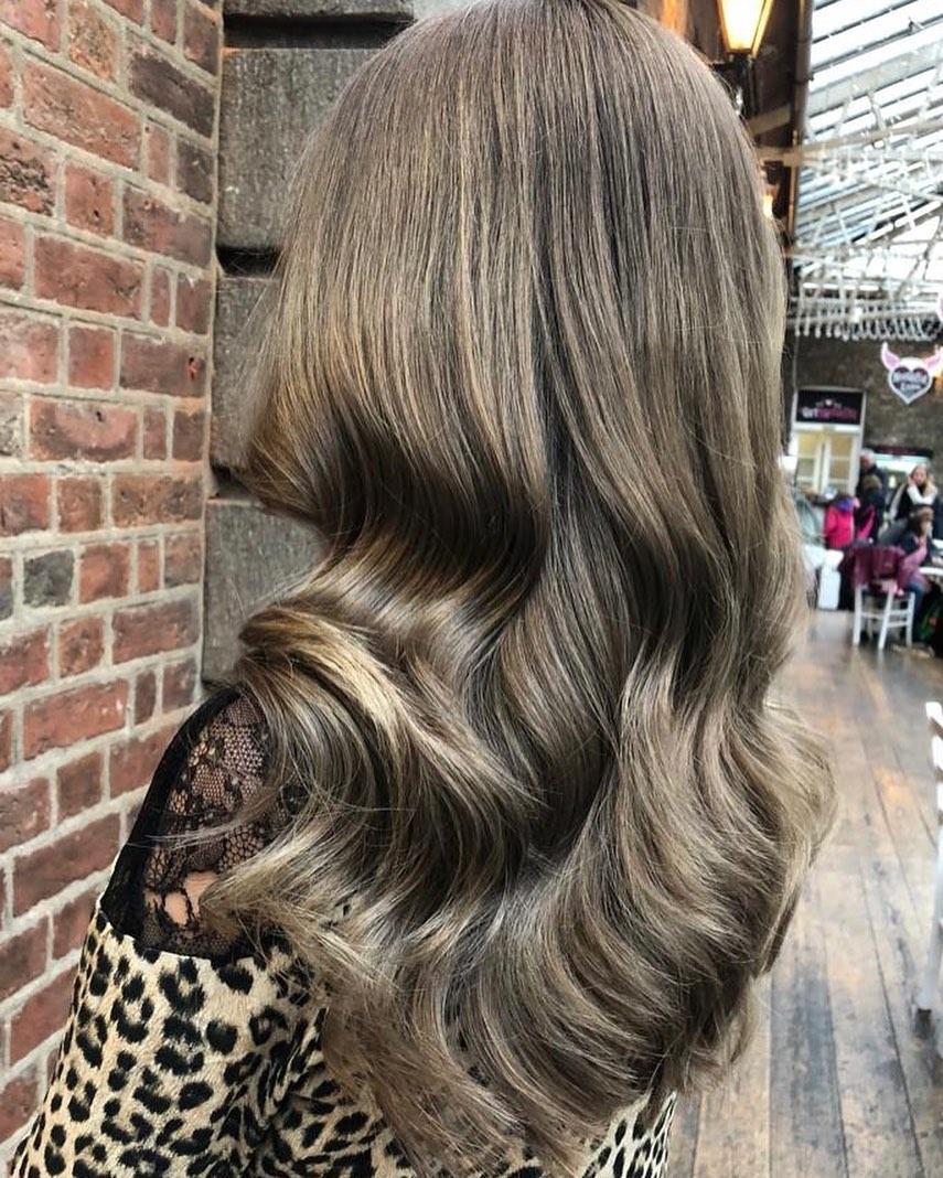 Back view of a woman with long, soft Hollywood waves in ash brown hair. The hair is glossy and voluminous, with smooth, flowing curls. She is wearing a leopard-print top with black lace shoulders and is photographed in an indoor market or arcade with brick walls and string lights.