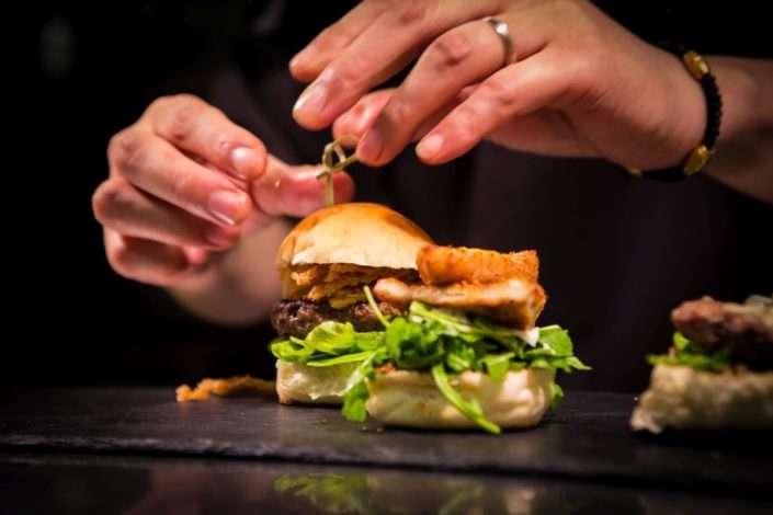 A person’s hands assembling a gourmet burger with arugula, crispy onion rings, and a toasted bun on a dark slate surface. A toothpick is being inserted to hold the burger together.