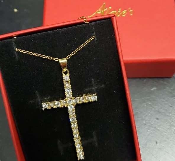 A gold chain necklace with a large cross pendant encrusted with sparkling rhinestones, displayed in a red gift box with black velvet lining. A partially visible red lid with gold script sits behind the box.