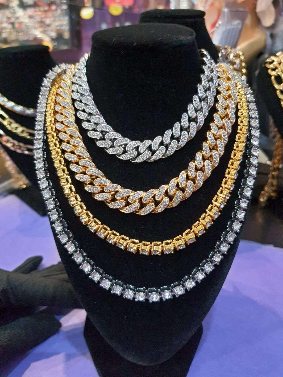 A display of multiple sparkling gold and silver chain necklaces on a black velvet bust. The necklaces include thick Cuban link chains and tennis chains, all encrusted with diamonds or rhinestones. Additional jewellery displays are visible in the background.