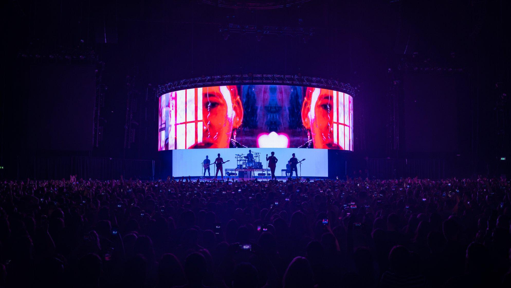 Silhouettes of a band performing on stage in front of a large LED screen displaying a vivid image of a woman’s face, with a crowd holding up phones in the darkened concert venue.