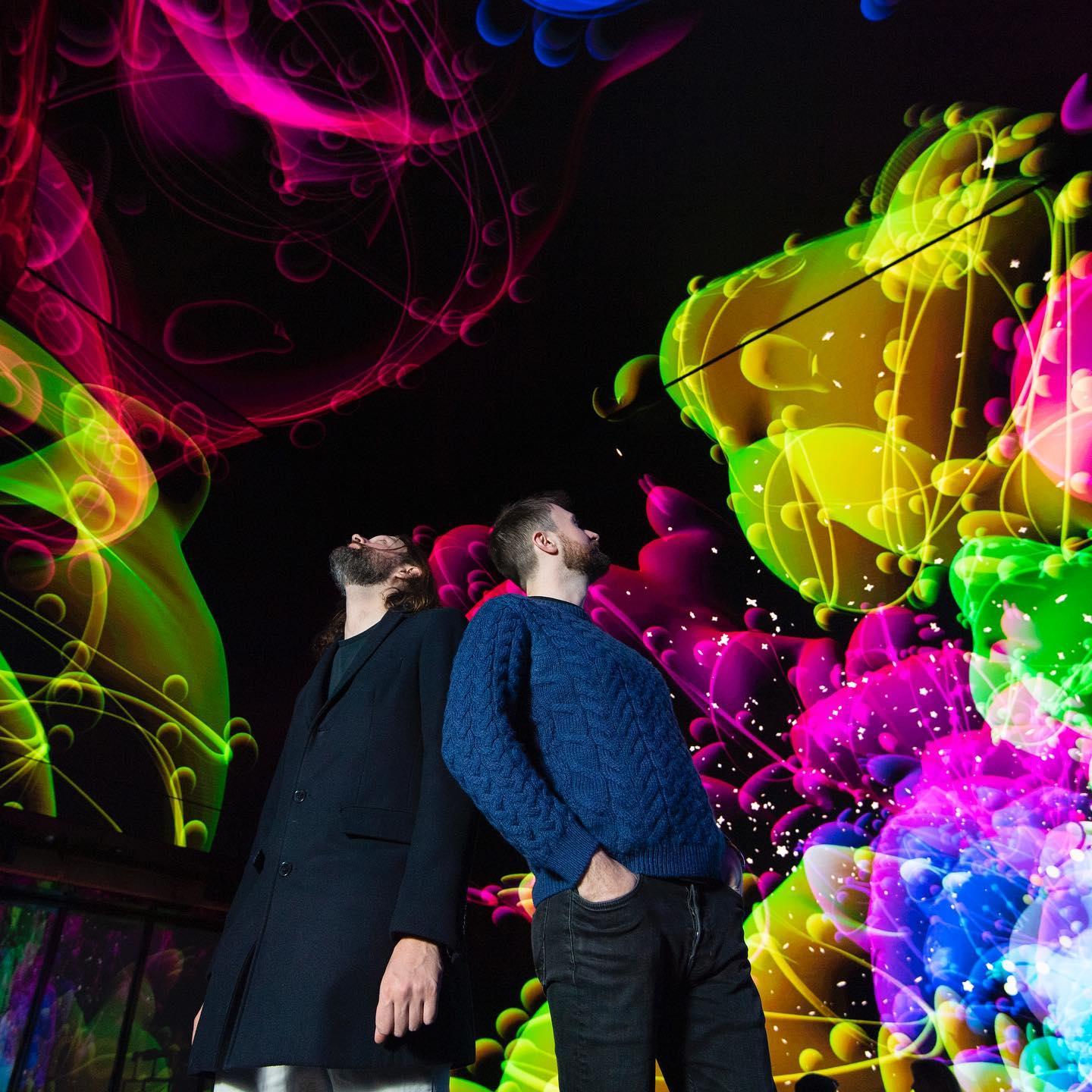 Two men stand under a swirling, colorful digital projection of neon-like pink, yellow, and green lights and shapes on a dark background, gazing upward.