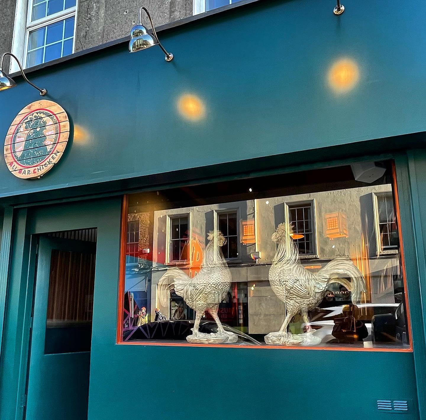 Exterior of a green storefront with a circular wooden sign featuring a stylized chicken. The large window displays two white rooster statues, with reflections of nearby buildings and street activity visible in the glass.