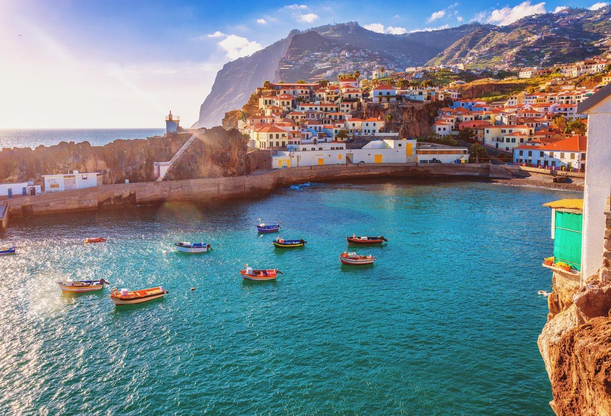 Small colorful fishing boats floating in a calm, turquoise harbor surrounded by a rocky breakwater. Beyond the harbor, a hillside village with tightly clustered houses featuring red-tiled roofs under a bright blue sky with scattered clouds.