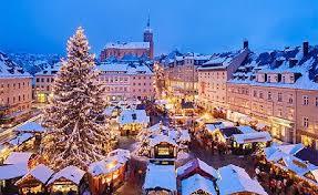 Snow-covered Christmas market at dusk with festive wooden stalls surrounding a large, decorated Christmas tree, and historic buildings illuminated in warm lights in the background.