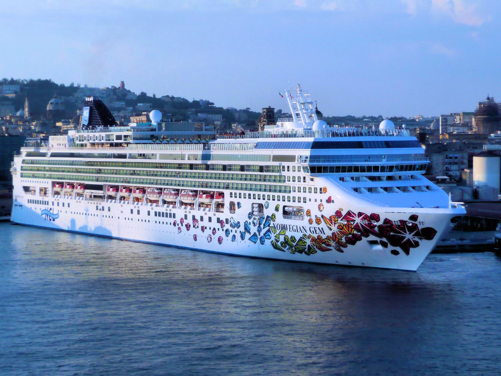 Large white cruise ship named Norwegian Gem with colorful geometric designs on its side, docked at a port with a cityscape in the background under a clear blue sky.