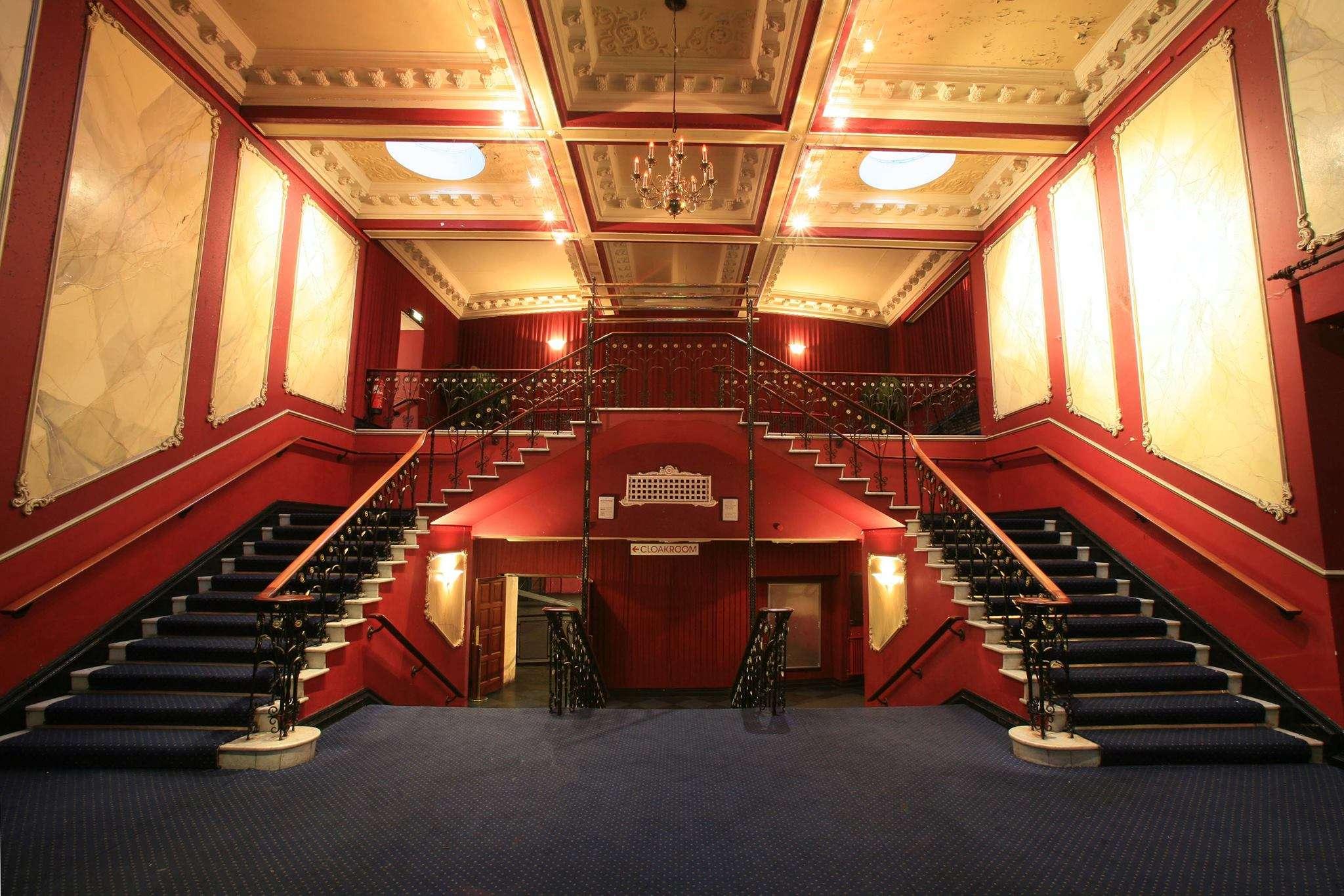 Symmetrical interior view of a grand staircase with red walls and ornate molding. The staircase splits into two flights leading upward with a blue carpet covering the steps and floor. Warm lighting from ceiling fixtures illuminates the space, and a sign pointing left to the cloakroom is visible above the central doorway.