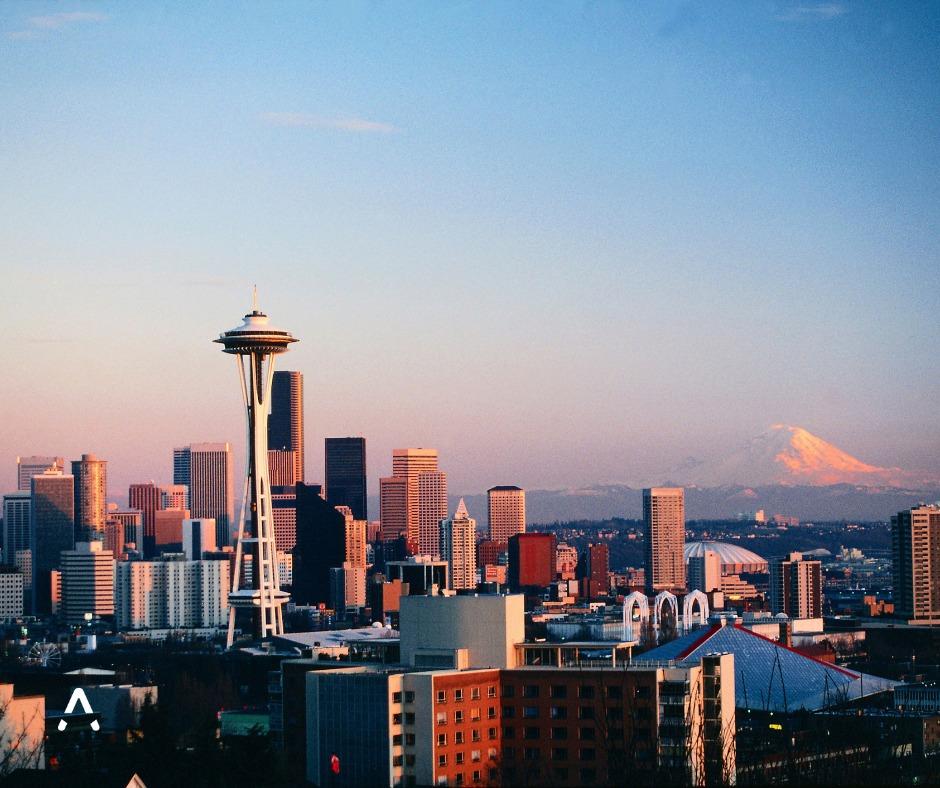 Cityscape of Seattle at sunset with the Space Needle in the foreground, modern high-rise buildings in the background, and Mount Rainier visible in the distance under a clear sky.