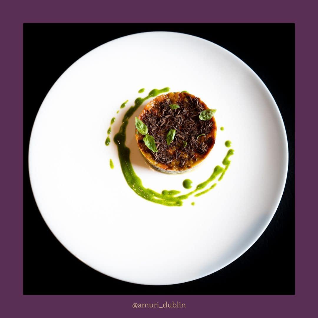 A round gourmet dish topped with finely chopped dark ingredients and small fresh basil leaves, presented on a large white plate with a decorative green sauce swirl around it, set against a black background.