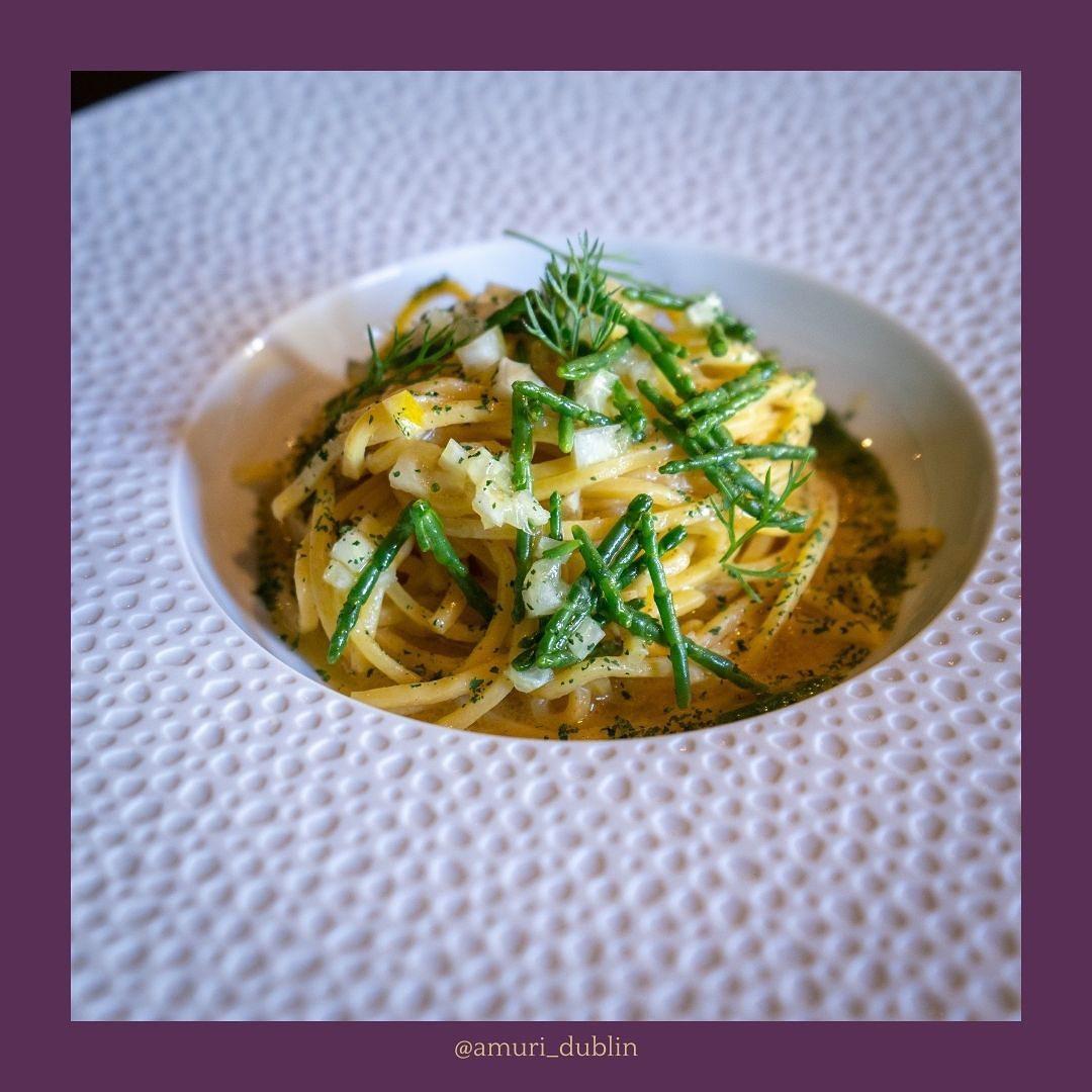 A neatly plated serving of spaghetti garnished with fresh herbs and green vegetables, placed in a textured white bowl on a patterned plate, with a purple border and the Instagram handle @amuri_dublin below.