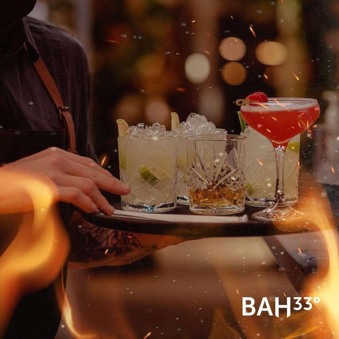A server carrying a tray with an assortment of cocktails, including a vibrant red drink garnished with a raspberry, crystal glasses filled with lime and ice, and a whiskey-style drink. The background is warmly lit with bokeh lights and decorative flame effects.