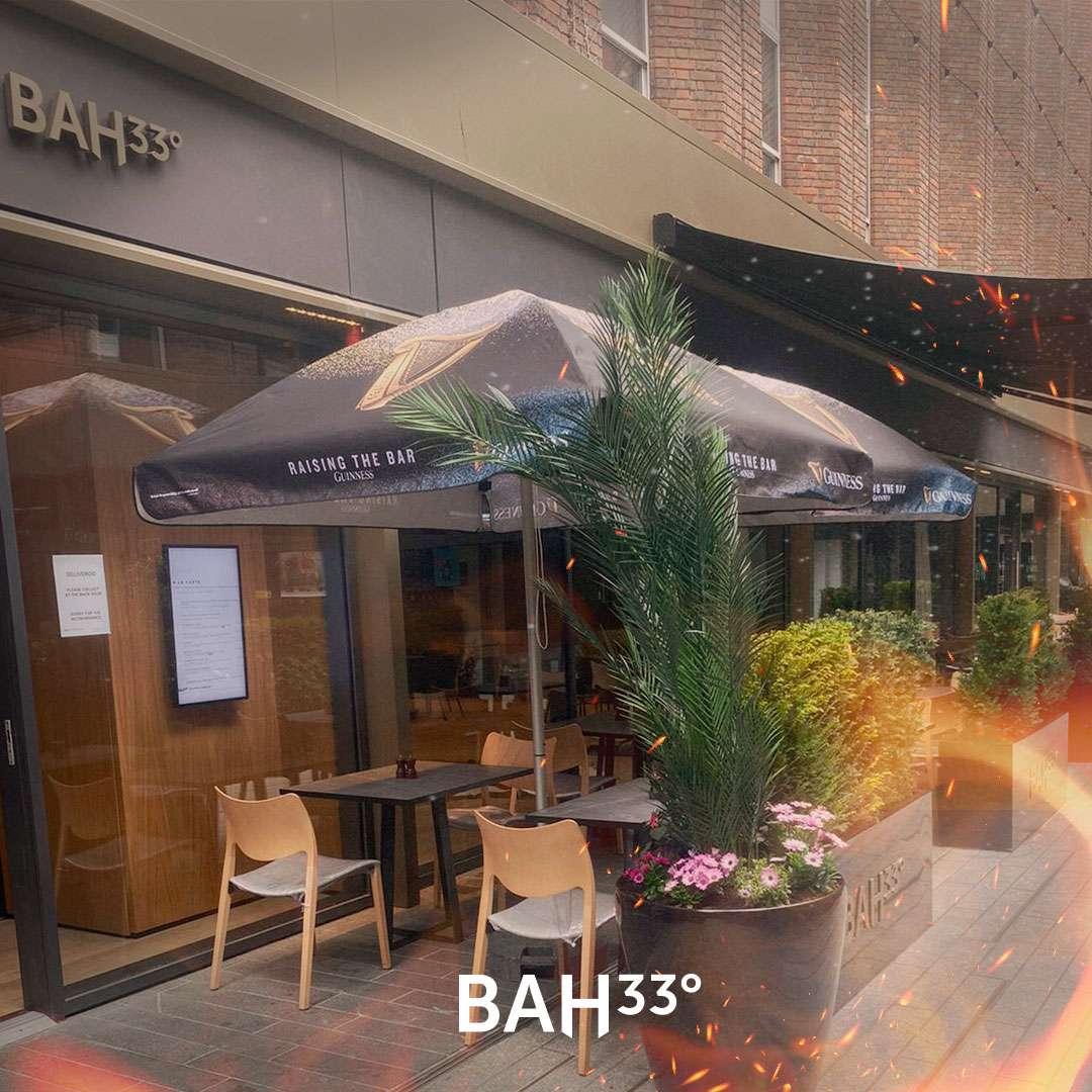 Street-level view of the BAH 33° restaurant exterior, featuring an outdoor dining area with modern tables and chairs under Guinness-branded umbrellas. Decorative planters with palm fronds and flowers line the sidewalk, creating a cozy and inviting atmosphere.
