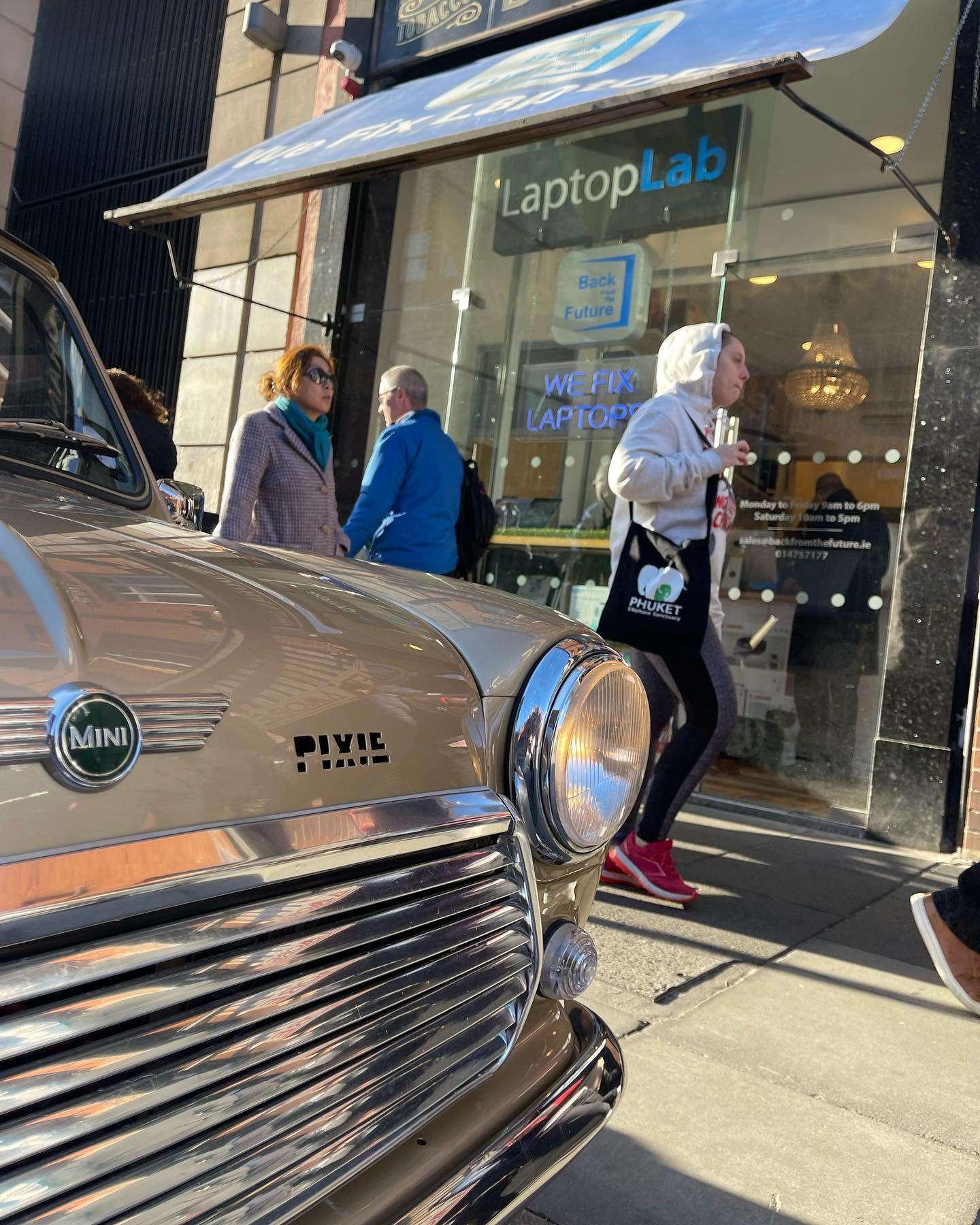 Front of a classic beige Mini car with "PIXIE" branding, parked in front of a LaptopLab repair store. Pedestrians walk by the shopfront, which displays signs reading "We Fix Laptops" and "Back from the Future."
