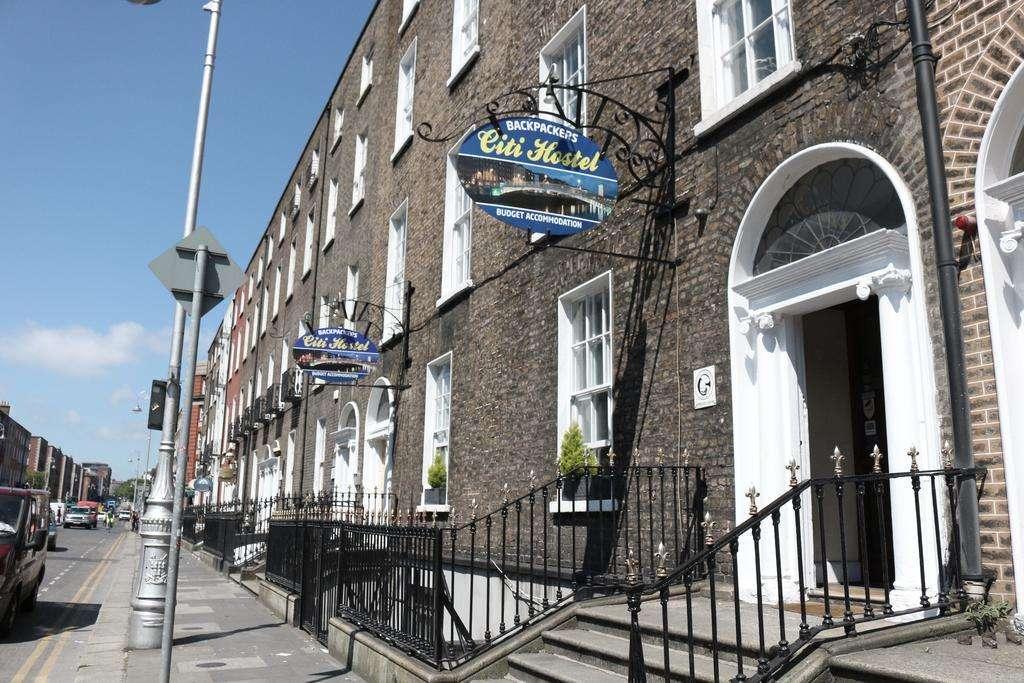 Row of Georgian-style brick buildings with arched doorways, housing the Citi Hostel, marked by blue hanging signs reading “Backpackers Citi Hostel – Budget Accommodation,” along a sunny city street.