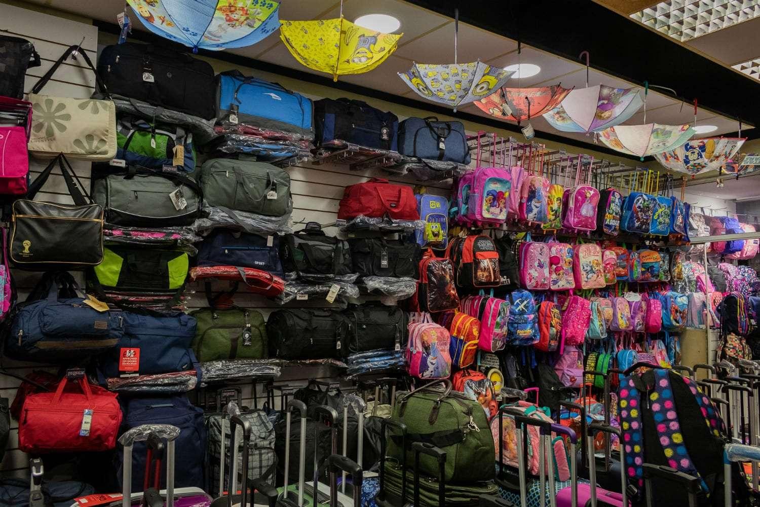 Store display with shelves of travel bags, backpacks in various colors and patterns, and children’s character-themed backpacks. Several open umbrellas hang upside-down from the ceiling, and rolling suitcases are lined up at the bottom.