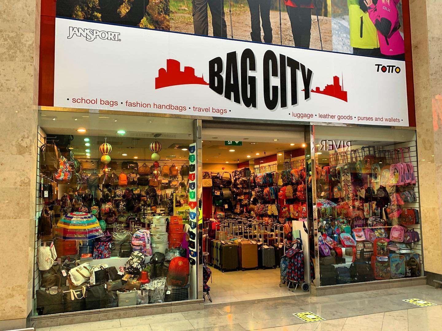 Storefront of Bag City in a shopping mall, showcasing a large selection of school bags, fashion handbags, travel bags, luggage, and backpacks displayed on shelves and racks behind glass windows