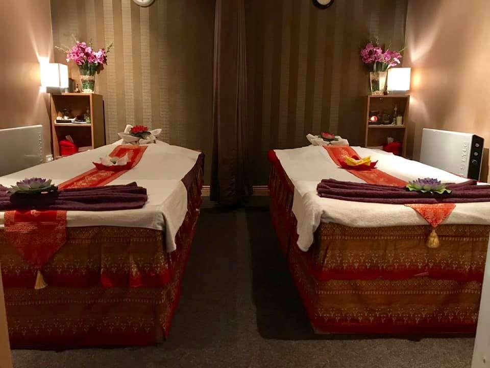 A cozy massage room with two massage tables side by side, each covered with white sheets and decorated with red and orange patterned runners. On each table, a dark purple towel is neatly folded with a decorative flower placed on top. The room has warm, ambient lighting from lamps on wooden shelves at the head of each table. The shelves hold pink flowers in vases and other small items. The walls are covered with brown striped wallpaper, and a curtain is drawn across the back wall.