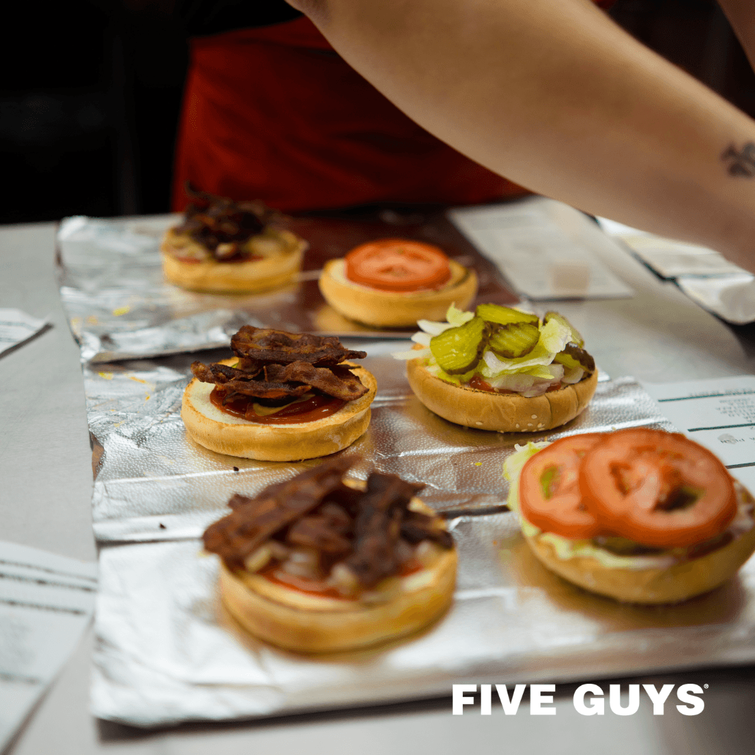 A behind-the-scenes view of burger preparation at Five Guys, showing several open-faced burgers laid out on sheets of foil. Each bun is topped with fresh ingredients like crispy bacon, pickles, lettuce, tomato, onions, and condiments, ready to be completed with patties and top buns. A crew member's arm reaches in from the right, actively assembling the burgers. The FIVE GUYS logo is prominently displayed in the bottom right corner.