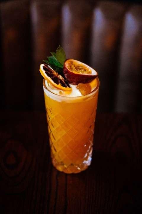 A tall glass of vibrant orange cocktail served on a dark wooden surface, garnished with a halved passion fruit, dried citrus slices, and a sprig of fresh mint. The glass has a diamond-cut pattern and is placed against a tufted leather background, creating an elegant and tropical presentation.