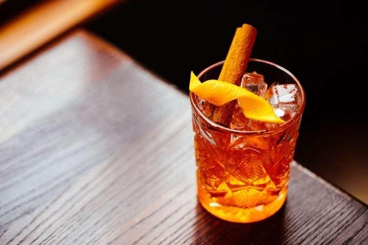 A glass of amber-colored cocktail served on a dark wooden surface, garnished with a cinnamon stick and a twist of orange peel, with several ice cubes inside the intricately patterned glass.