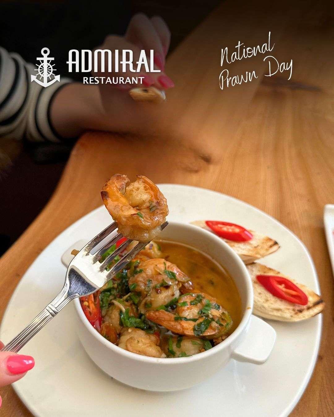 Fork lifting a garlic prawn over a bowl filled with prawns in herb and oil sauce, served with toasted bread topped with red chili slices. “Admiral Restaurant” and “National Prawn Day” text is displayed on the image.