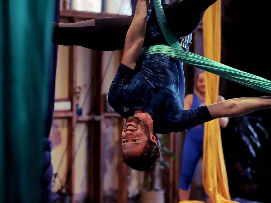 Man hanging upside down in a green aerial silk hammock during an aerial yoga or acrobatics class, smiling with arms extended. A second person in the background prepares with yellow silks in a studio space.