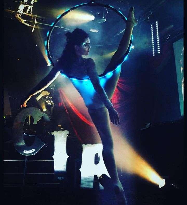 A female aerial performer in a shimmering outfit and face makeup poses inside a glowing aerial hoop, balancing gracefully with one leg extended high. Stage lights and theatrical smoke create a dramatic effect, with large decorative letters spelling "CIR" in the background.