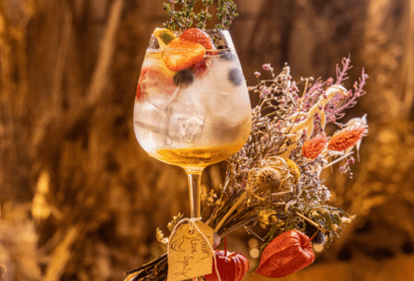 Elegant cocktail in a stemmed glass garnished with fresh berries, citrus slices, and a sprig of thyme, placed next to a decorative bouquet of dried flowers.