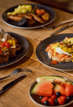 Variety of colorful breakfast dishes including smoked salmon with poached eggs, a fruit platter with watermelon and berries, a traditional English breakfast, and a pastry topped with berries and edible flowers.