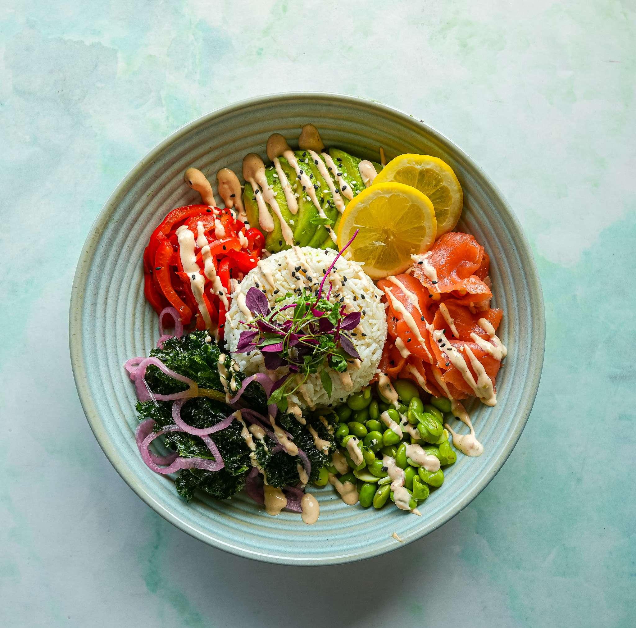 Colorful poke bowl with sliced salmon, avocado, red pepper, lemon, rice, kale, edamame, pickled onions, and drizzle of dressing in a blue bowl.