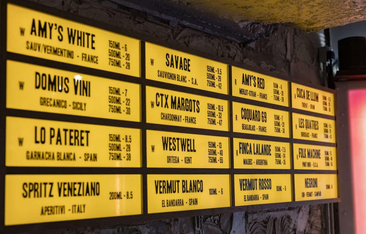 A backlit yellow menu board mounted on a wall, listing various wines and aperitifs along with volume and prices, such as Amy's White, Savage, and Domus Vini.